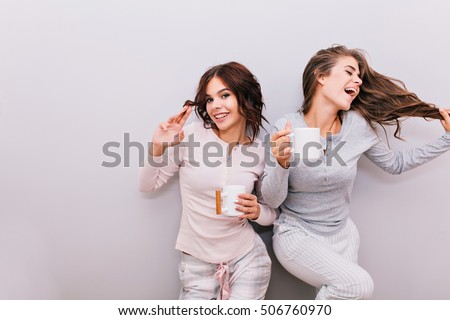 Two beautiful girls in pajamas having fun on gray wall background. Girl with long hair laughing and keeps eyes closed, other with curly hair smiling to camera
