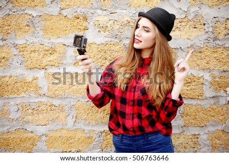  Hipster teenage girl in black hat, red plaid shirt taking a selfie on action camera against brick wall. 