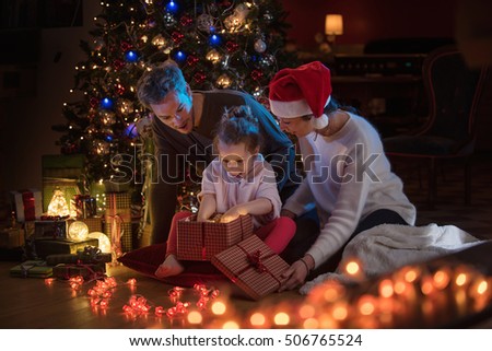 Christmas night. Near the christmas tree a lovely family opening their gifts. They enjoy the warm Christmas atmosphere in their living room, mom is wearing a hat of Santa Claus.