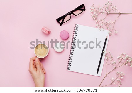 Woman hand hold cup of coffee, cake macaron, clean notebook, eyeglasses and flower on pink table from above. Female working desk. Cozy breakfast. Flat lay style.