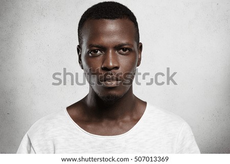 Close up portrait of good-looking serious African man with healthy clean skin wearing white casual t-shirt posing isolated against gray studio wall with copy space for your promotional content