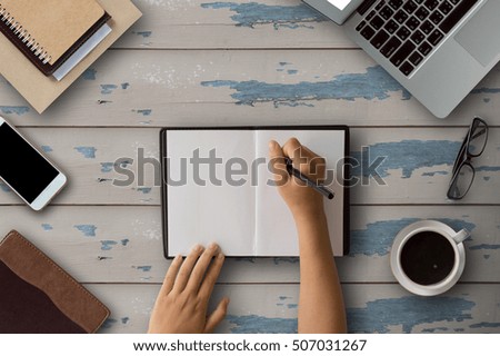 Desktop wooden office with laptop computer, note pad, smart phone,glasses and coffee of cup.Female hands Write message in notebook on wood background..Coffee cup on  wood background.Copy space.