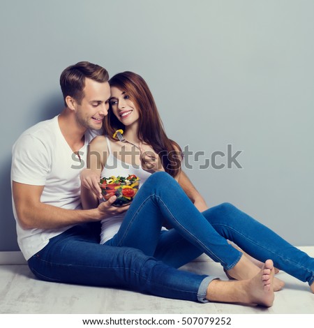 Beautiful young playful couple eating salad together, sitting on floor. Caucasian models in vegetarian, weight lossing, dieting, healthy food concept studio shot, against grey background.