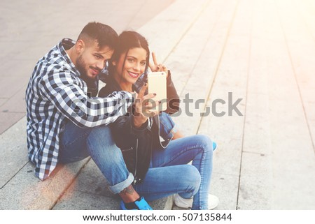 Stylish young friends are sitting on the steps and having fun during recreation time. They taking a selfie on mobile telephone and smiling at the camera. Cute couple look really happy during vacations