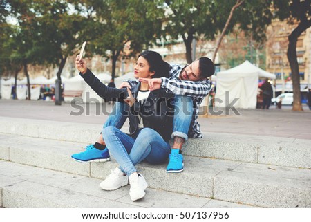 Beautiful hipster girl and a handsome young guy are sitting on the steps outside and taking a selfie on the white smartphone at weekend. Happy romantic couple shooting video during recreation time