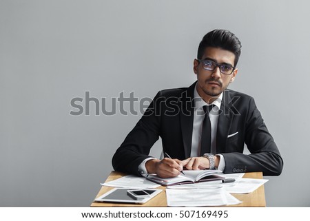 Businessman writing in a notebook and looking at camera  on a grey background, the tablet, telephone and papers on the table. copy space