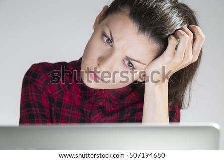 Close up of serious young woman in red dress sitting in her room and looking at her laptop screen. Concept of pondering