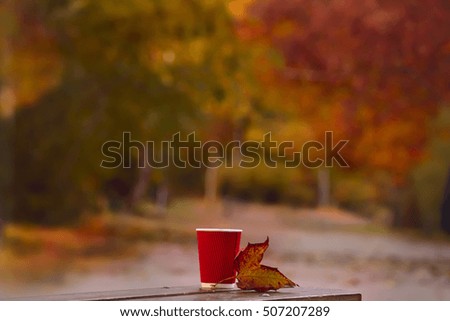 paper and a glass of red autumn leaf on a bench next. In the background is blurred image naturalnnnogo autumn park autumn. Autumn red and orange colors of autumn leaves and trees.
