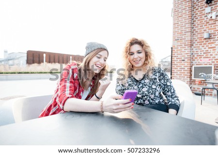 Two young curly and straight blonde hair caucasian woman sitting on a bar, using smartphone, both looking downward and tapping the screen - technology, social network, communication concept