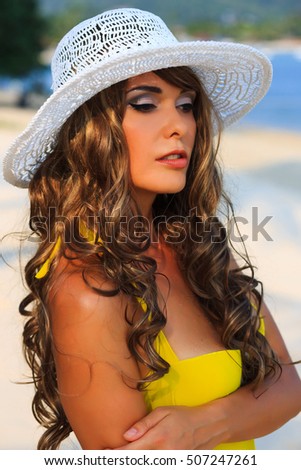 Young beautiful smiling woman in hat and yellow bikini is posing on the beach