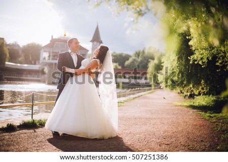 in love with a beautiful young bridal couple kissing near lake in a sunny day. 
bride and groom dancing on the pier under tree. backlight of the sun. Exciting honeymoon photo/ picture. Copy space.