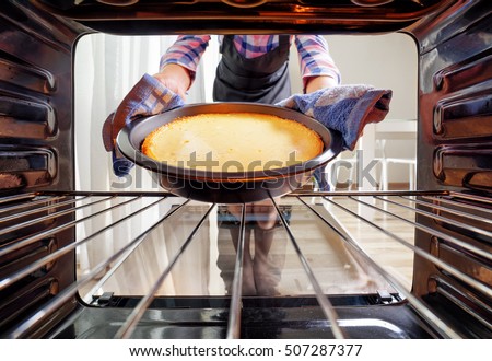 Housewife using dishcloth for taking cheesecake out of oven in kitchen. View from inside of the oven. Woman wearing colorful checkered shirt and black apron. Royalty-Free Stock Photo #507287377