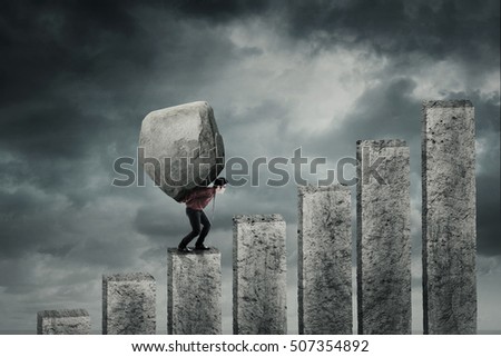 Young businessman walking on the financial chart while carrying a big stone. Concept of hard work Royalty-Free Stock Photo #507354892