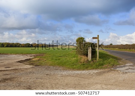 a wooden footpath signpost near a country highway with colorful woods in a yorkshire wolds landscape in autumn under a blue cloudy sky