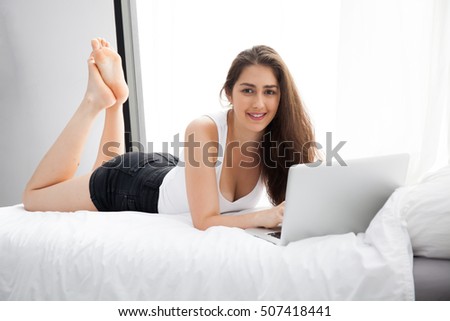Beautiful Caucasian woman laying down on white bed using laptop and smile