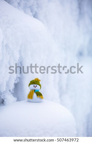 Nice dreaming snowman in green hat is standing on interesting textured snow near to the hill on cold winter day.