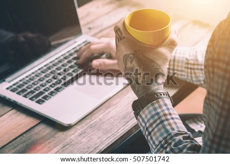 Male Hands With Tattoo Holding Yellow Cup And Typing On The Keyboard Of A Laptop On Vintage Wooden Table. Mock-up With Laptop