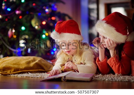 Two cute little sisters reading a story book together under a Christmas tree on Christmas eve at home
