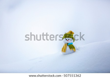 Nice dreaming snowman in green hat is standing on interesting textured snow near to the hill on cold winter day.