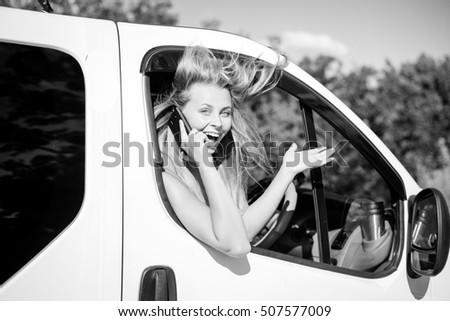 Black and white picture of joyful blond girl speaking phone and looking from car window. Young woman excited and smiling while talking with someone on sunny countryside background.