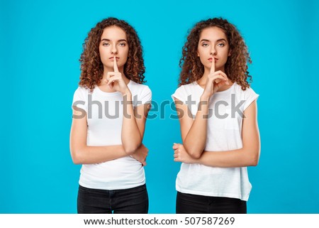 Two pretty girls twins showing keep silence over blue background.