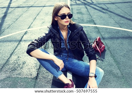 young stylish hipster woman outdoor, wearing sunglasses, black leather jacket, jeans overall, suede sneakers, leather backpack, urban fashion