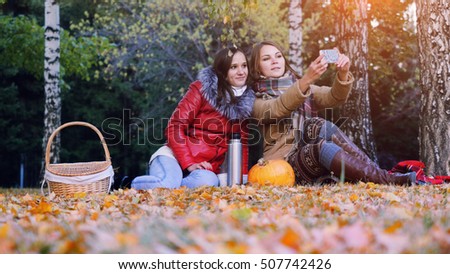 Girls making selfie on a picnic in autumn park sitting  the fallen leaves near the pumpkin at halloween time.