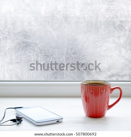 Cup of coffee, smartphone and headphones on a windowsill. In the background frosty pattern on window as a Christmas background