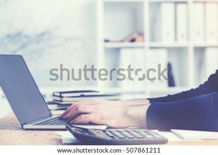 Side view of accountant's hands using laptop with blank screen on cork desktop with calculator and paperwork. Mock up. Toned image