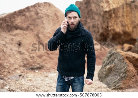Close up portrait strong man speaking by phone.fisherman on the shore.Street outfit,azure hat,black pullover,denim,man and rocks,bearded man