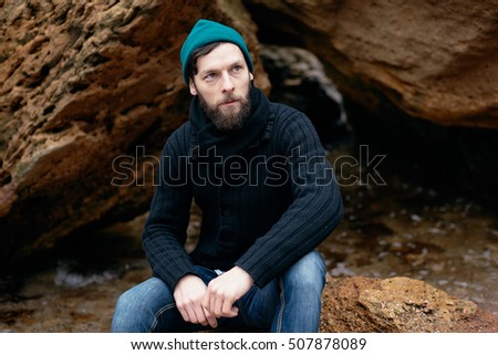 Close up portrait strong man fisherman on the shore.details, sailor hat,black pullover, denim,alone man with rocks,man sitting on the sea.street outfit.