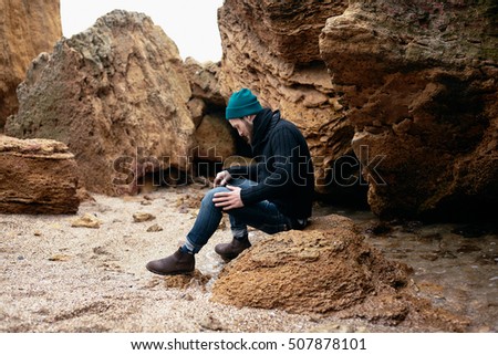 Close up portrait strong man sitting on the stone.fisherman on the shore.jeans,casual clothes,hat,black pullover,brow boots,shoes store,yellow stones,fashion style,trend