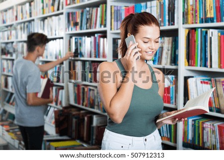 Cheerful teenager girl holding open book and talking on mobile phone in shop