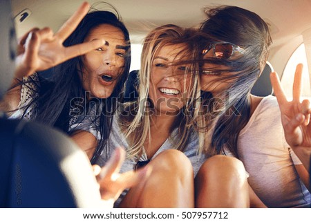 Three beautiful female friends make peace signs while seated in back seat of car with wind in their hair