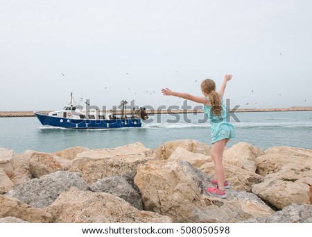 Eight year old girl standing on the rocks of the Denia harbour wall, or breakwater and waving at an incoming fishing boat being circled by seagulls