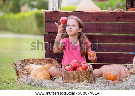 Young girl holding apple and sitting on hay among pumpkins in the garden