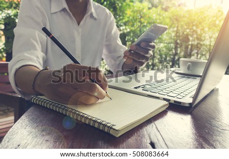 Woman write on notebook and holding mobile phone with laptop vintage tone lens flare effect.