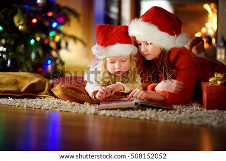 Two cute little sisters reading a story book together under a Christmas tree on Christmas eve at home