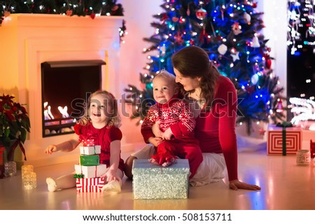 Family on Christmas eve at fireplace. Mother and little kids opening Xmas presents. Children with gift boxes. Living room with traditional fire place and decorated tree. Cozy winter evening at home.