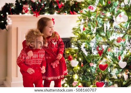Happy little kids in matching red knitted sweaters decorate Christmas tree in beautiful living room with traditional fire place. Children opening presents on Xmas eve.