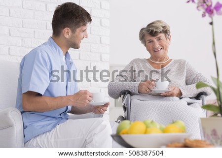 Elder woman sitting on a wheelchair with cup of tea, close to her male nurse
