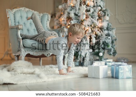 cheerful blond boy with blue eyes in gray shirt and light trousers slide with blue chair standing next to a Christmas tree stretching for gifts. new Year. Christmas
