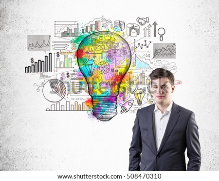 Portrait of young businessman standing near colorful light bulb sketch surrounded by graphs on concrete wall