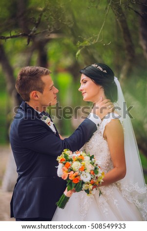 Elegant stylish groom with his happy gorgeous brunette bride on the background of trees in the park. Kissing picture.