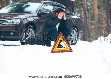 Angry young man waiting for help, sitting near the broken car on the side of the road in the winter in the woods