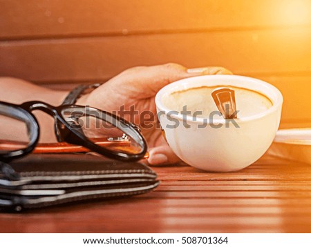 Eyeglasses resting on a leather wallet and made with orange light color filters and blurred man holding white coffee cup background.