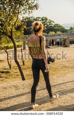 Young tourist in photo walk with professional digital camera hand on shoulder against summer park. back rear view. 