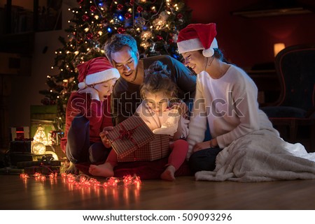Christmas night. Near the christmas tree a lovely family opening their gifts. They enjoy the warm Christmas atmosphere in their living room, mom and kids wearing a hat of Santa Claus.