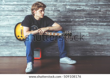 Handsome young guy with electric guitar sitting on amplifier in wooden room. Music, concert rehearsal concept