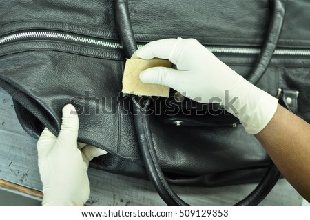 Black Leather Bag cleaning with a Microfiber cloth in a leather factory. The hands of the worker covered with gloves are into sharp focus and no human face is visible in the image.  Royalty-Free Stock Photo #509129353